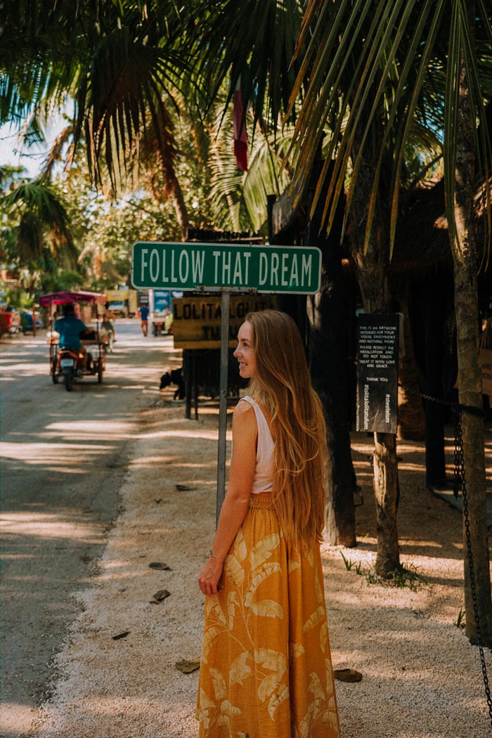 Travel blogger on a beach road in front of "Follow that Dream" sign in Tulum, Mexico.